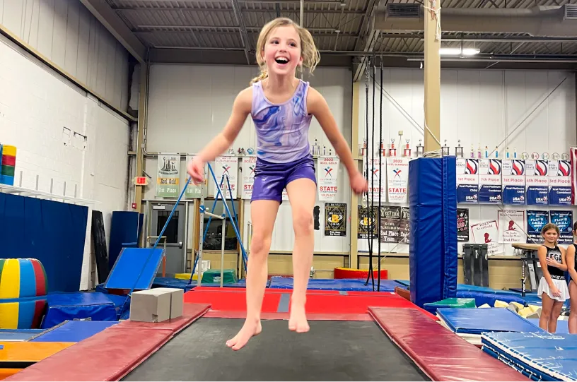 This image shows a young girl jumping on a gymnastics power launch mat in a training gym at Excel Gymnastics Academy.
