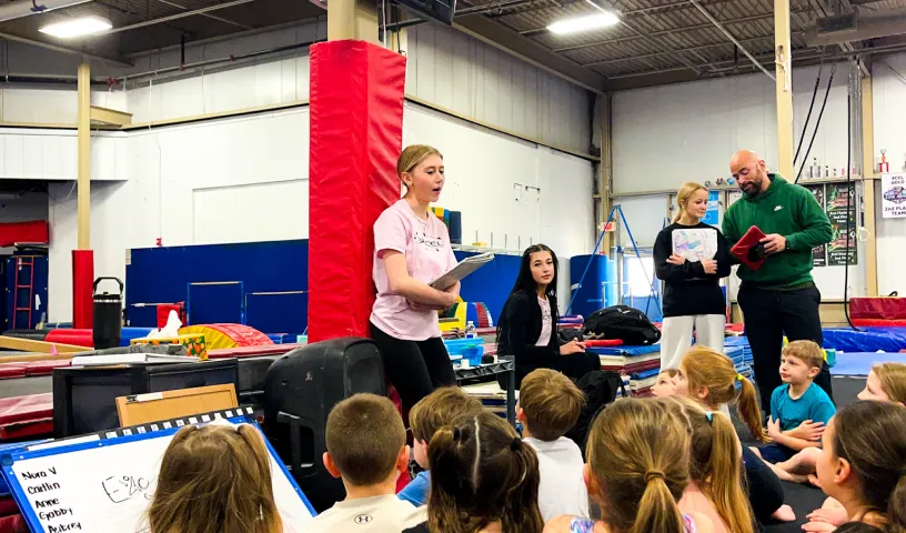 This Image shows a coach or instructor is speaking to a group of young children seated on the floor at Excel Gymnastics Academy.