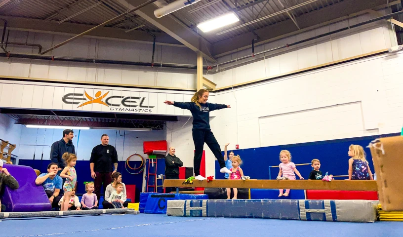 This image shows a young gymnast is practicing balance on a beam while other children watch at Excel Gymnastics Academy