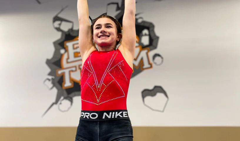 The image shows a young girl wearing a red decorated gymnastics leotard and black Nike Pro athletic shorts.