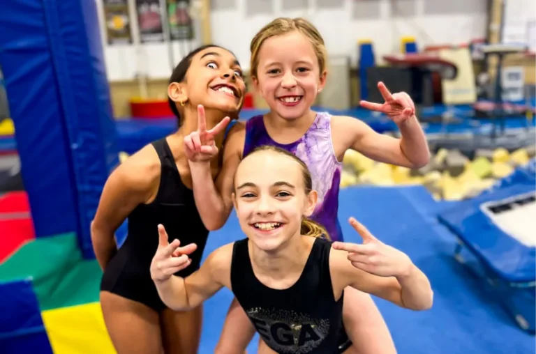 This image shows three young gymnasts smiling and posing for a picture inside a training gym at Excel Gymnastics Academy